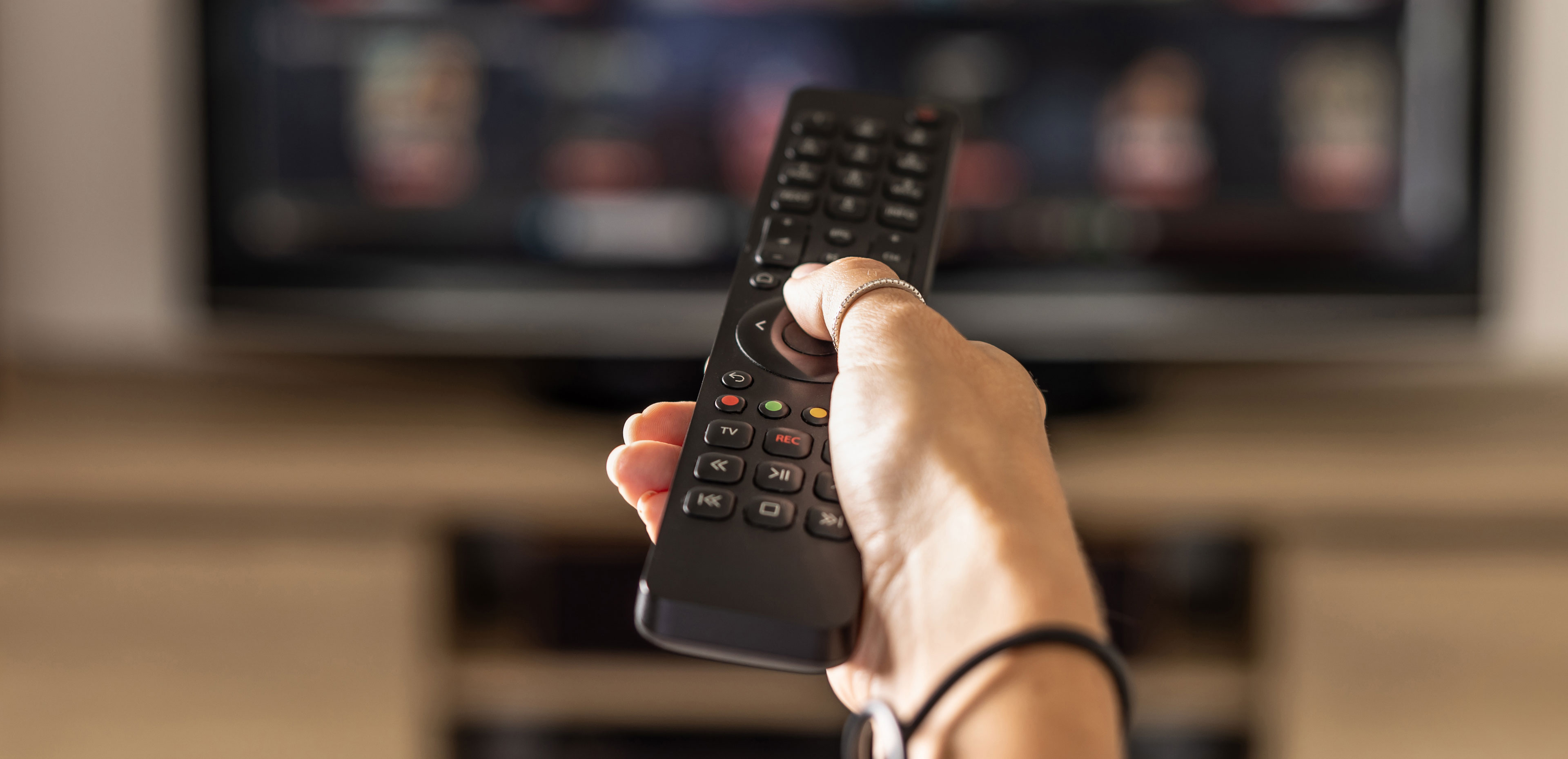 Hand holding a TV remote toward a television, representing the growing complexity of finding and watching live sports across multiple streaming services.